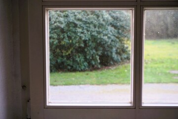 Innenausstattung mit weißem Holzrahmenfenster und Ausblick auf Kieselweg, grüner Wiese mit grünem Busch bei Nebel am Morgen im Winter