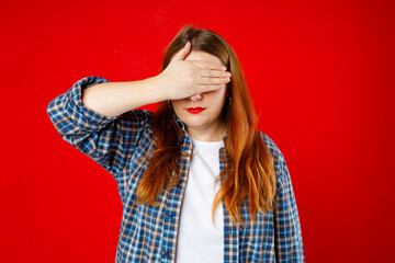 Portrait of young scared woman covering eyes with one hand while standing against red background....