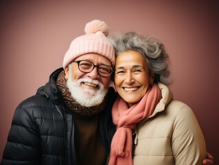 Elderly multiracial couple on pink background. A man in a knitted cap with a pump and a down jacket and a cute lady in a beautiful scarf. couple smiling and posing for the camera.