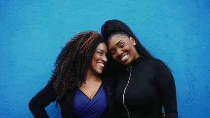 Happy African American mother and adolescent girl posing together in bonding embrace, authentic genuine love and affection between parent and offspring in front of blue wall