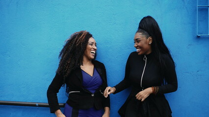 Happy mother and adolescent daughter leaning on blue wall in urban street conversing and laughing...