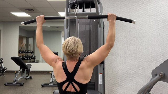 An adult Caucasian woman trains pull-ups with a horizontal bar on a machine in the gym. Rear view, horizontal video. The concept of a sports lifestyle in adulthood. Woman on simulator, back view.