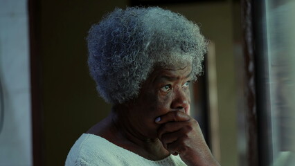 One contemplative senior black 80s woman standing at home with hand on chin pondering deeply about...