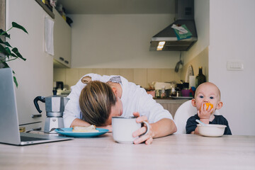 Obraz na płótnie Canvas Young mother with coffee fighting tiredness while breakfast with baby in kitchen. Freelancer mom and child after sleepless night. Woman studying or working online at home on maternity leave