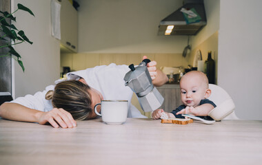 Exhausted young woman with baby is sitting with coffee in kitchen. Modern tired mom and little...