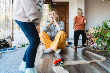 Stressed out mother sitting in middle of toys while children naughty running around her at room.