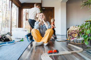 Stressed out mother sitting in middle of toys while children naughty running around her at room. - obrazy, fototapety, plakaty
