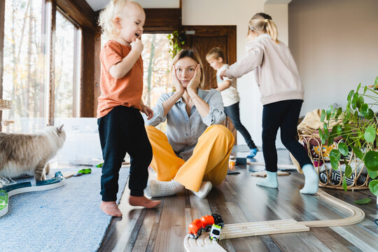 Stressed out mother sitting in middle of toys while children naughty running around her at room.
