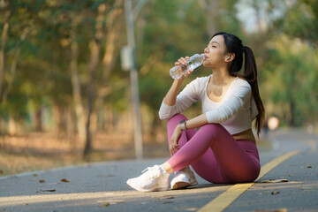 Fototapeta premium Asian woman drinking water from a bottle after running with a public park background, outdoor workout concept, drinking clear mineral water after jogging.