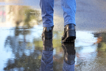Female legs in boots on a street with puddles. Woman reflected in water, walking in spring weather