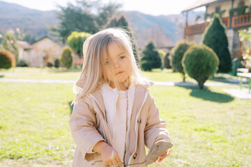 Little girl with a dry leaf in her hands stands with her head tilted to the side on a green lawn