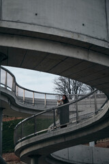 Fototapeta premium A young woman poses with a dog standing on a beautiful architectural bridge in the center of Belgrade on the embankment. Female owner with red welsh corgi Pembroke, urban lifestyle.