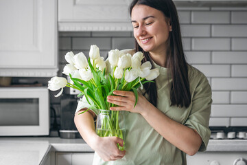 A woman holds a vase with white tulips in a kitchen interior.