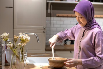 Pretty muslim Asian woman in hijab wearing a purple dress, making dough in a modern kitchen with...