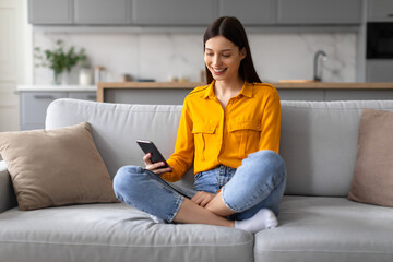 Smiling young woman browsing on phone while sitting on sofa