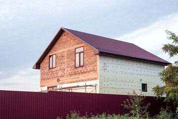 A two-story house under construction with a corrugated roof in the countryside. Insulation of house walls with polyurethane