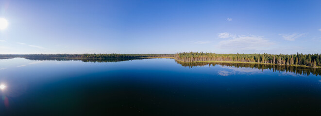 Panoramic aerial view of a large calm northern lake reflecting a clear blue sky.  The lakeshore is heavily treed with spruce and the sun reflects in the water on the left side of the image.
