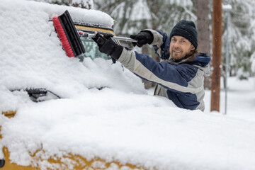 A man is standing in the snow, holding a snow brush and smiling. He is cleaning the windshield of a yellow jeep