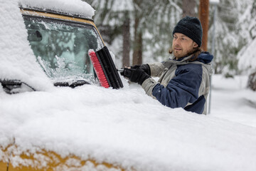A man is cleaning his car's windshield with a snow brush. The scene is set in a snowy environment, and the man is in a good mood