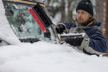A man is cleaning the windshield of a car with a snow brush. The scene is set in a snowy...