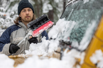 A man is clearing snow off a car with a snow brush. The scene is set in a snowy environment, and...