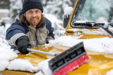 A man is smiling while he is shoveling snow off of a yellow vehicle. The scene is set in a snowy...