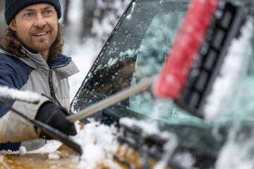 A man is cleaning his car's windshield with a snow brush. He is smiling and he is enjoying the task