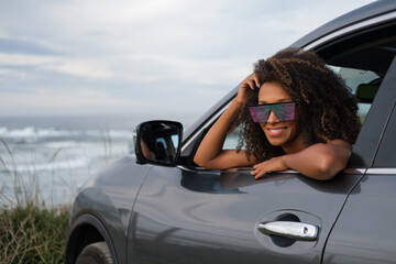 Portrait of a young black woman wearing sunglasses, leaning out of her car window during a relaxing getaway to the coast. - 751568815