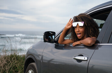 Portrait of a young black girl with an funny expression of wonder as she stands out through her car window with the sea and sky in the background. - 751568823