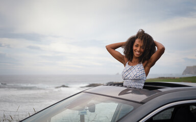 Blissful young black woman leaning out of the sunroof of her car with the sea and sky in the background. Female driver enjoying the freedom of a getaway.