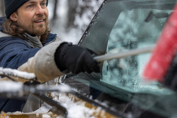 A man is cleaning his car's windshield with a snow brush. The man is smiling and he is enjoying the task