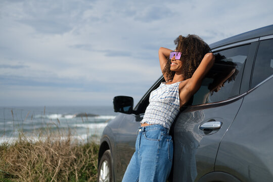 Relaxed and blissful young black woman leaning on her car in front of the sea. Getaway to take a break and enjoy the outdoors and freedom.