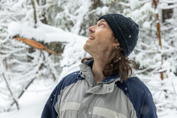 A man wearing a black hat and a blue and gray jacket is standing in the snow. He is smiling and looking up at the sky