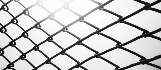 Fototapeta premium A black chain link fence stands starkly against a white sky background. The intricate pattern of the fence creates a striking visual contrast.