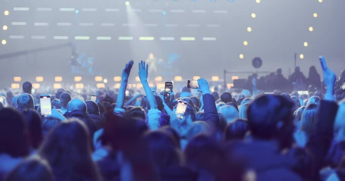 Back view, happy people crowd on blurred rock band stage background in blue spotlights light. People move, dance, wave arms, film performance of favorite musical group on phones. Concert entertainment