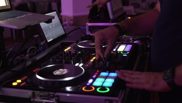 Frame in motion on the side with the hands of a DJ mixing at his desk, and turning the knobs while dancing. Colored lights playing in the ballroom