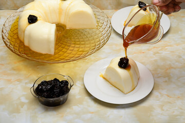 Blancmange decorated with plums, syrup falling over a slice on a white plate, yellow marble bottom