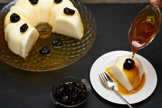 Blancmange decorated with plums, syrup falling over a slice on a white plate, black background.