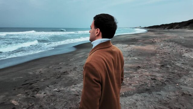 Fascinating Italian model actor looks at the sea well dressed ready for the cinema scene during the production a movie walks on the beach on a gray day and smiles looking at the video camera.