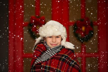 Funny boy with a fur hat and wrapped in a plaid plaid against the background of falling snow. The child is waiting for Christmas gifts.