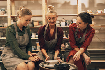 A company of three pretty young women friends make ceramic mugs in a pottery workshop.
