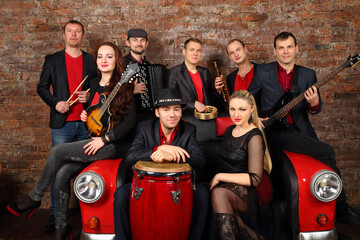 Brass band of eight people in red and black clothes pose in studio with car couch