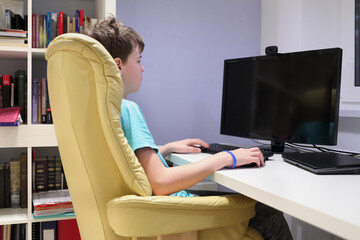 Teen boy sitting in chair at computer in his room and looking at monitor, side view