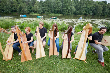 Ensemble of seven musicians play celtic harps performing outdoors at pond shore