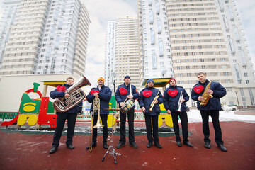 Brass band of six musicians pose on playground near buildings at winter day