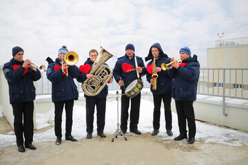 Brass band of six musicians play on roof of tall building at winter