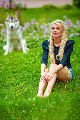 Young blond woman sits on grassy lawn in park, dog sits behind her out of focus