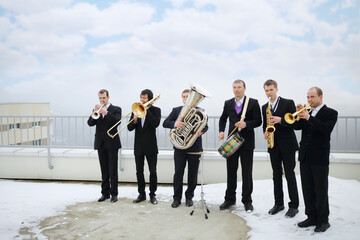 Brass band of six musicians in suits play on roof of tall building at winter
