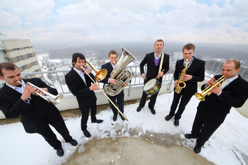 Brass band of six musicians in suits play music on roof of tall building at winter