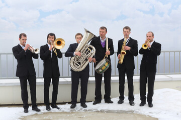 Brass band of six musicians in suits play music on roof of tall building at winter day